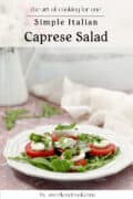 A plate of Caprese salad with sliced tomatoes, mozzarella, arugula, basil, and balsamic glaze, set on a table with a light cloth and a vase in the background.