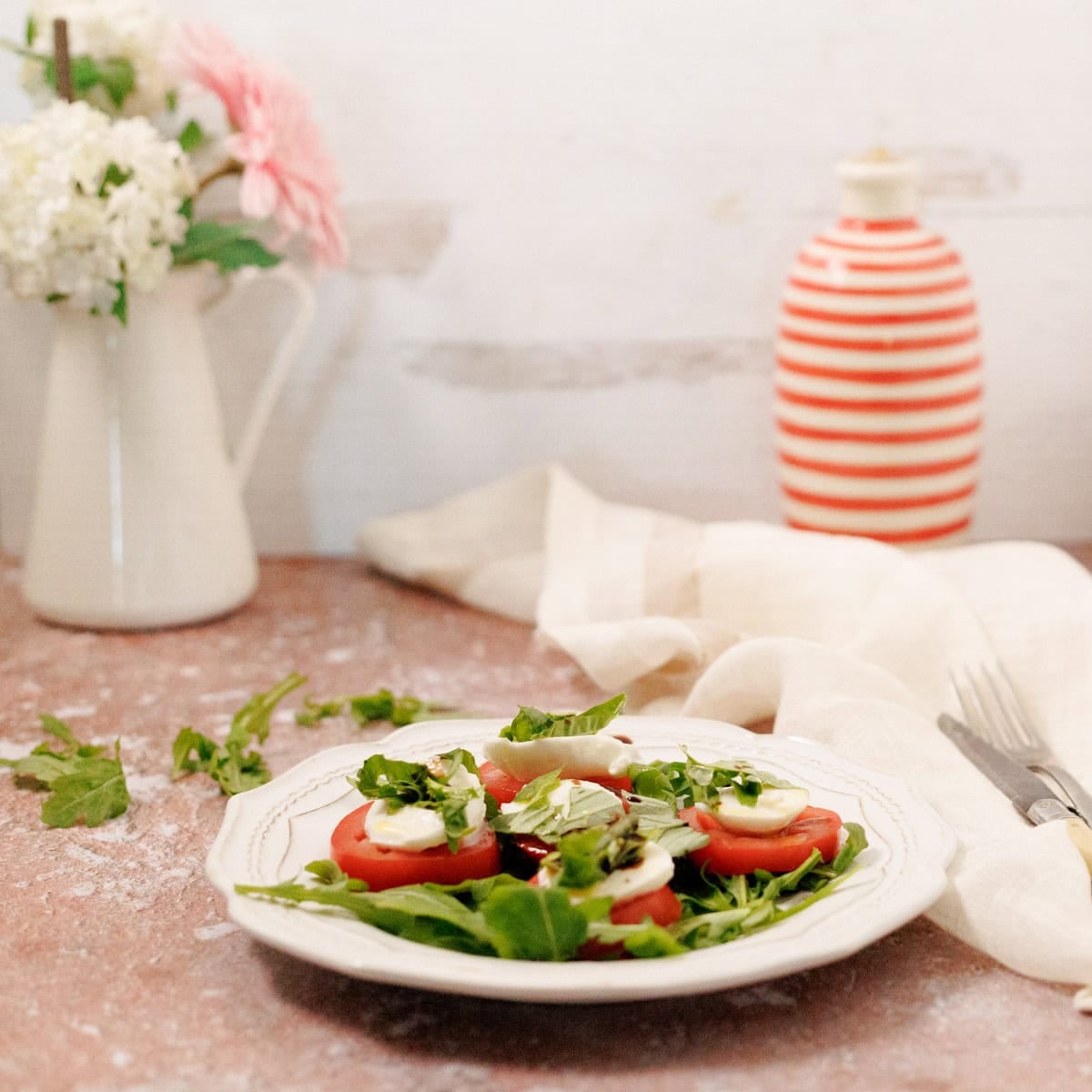 A plate of sliced tomatoes and mozzarella on arugula sits on a table with a white napkin, cutlery, a striped bottle, and a vase of flowers in the background.