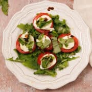 A plate of caprese salad with sliced tomatoes, mozzarella, fresh basil, arugula, and a drizzle of balsamic glaze on a white plate.