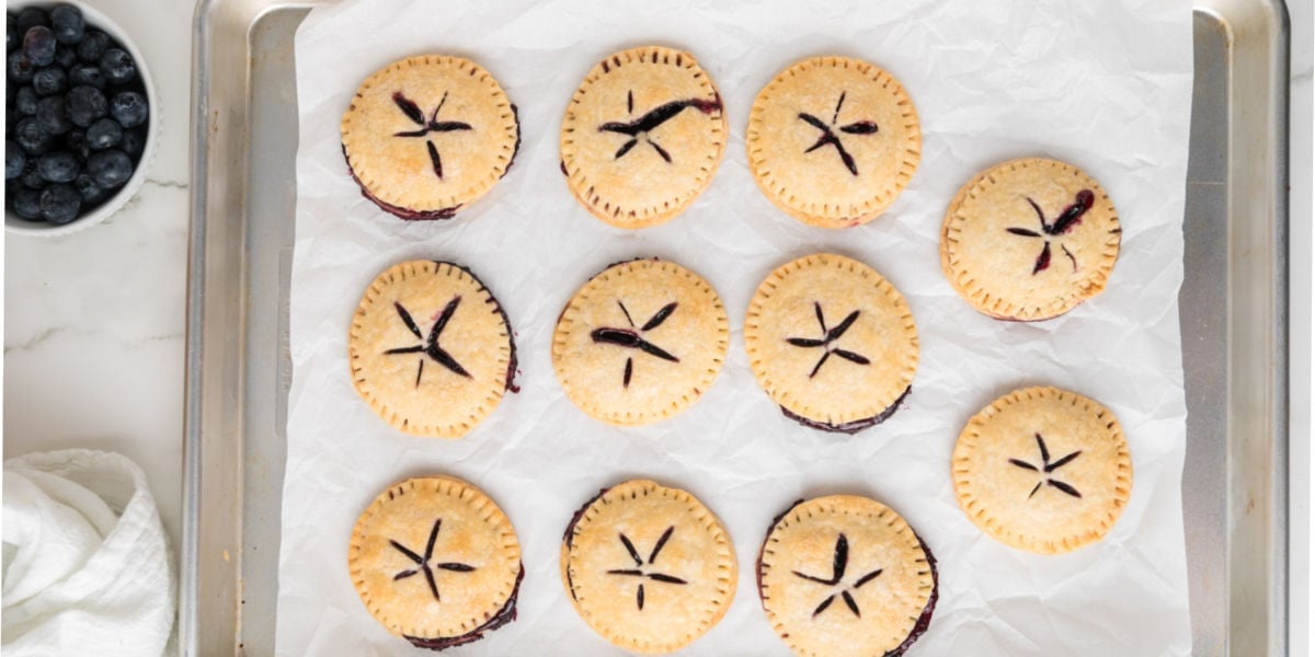 A baking sheet lined with parchment paper holds twelve baked mini blueberry hand pies, with a bowl of fresh blueberries nearby.