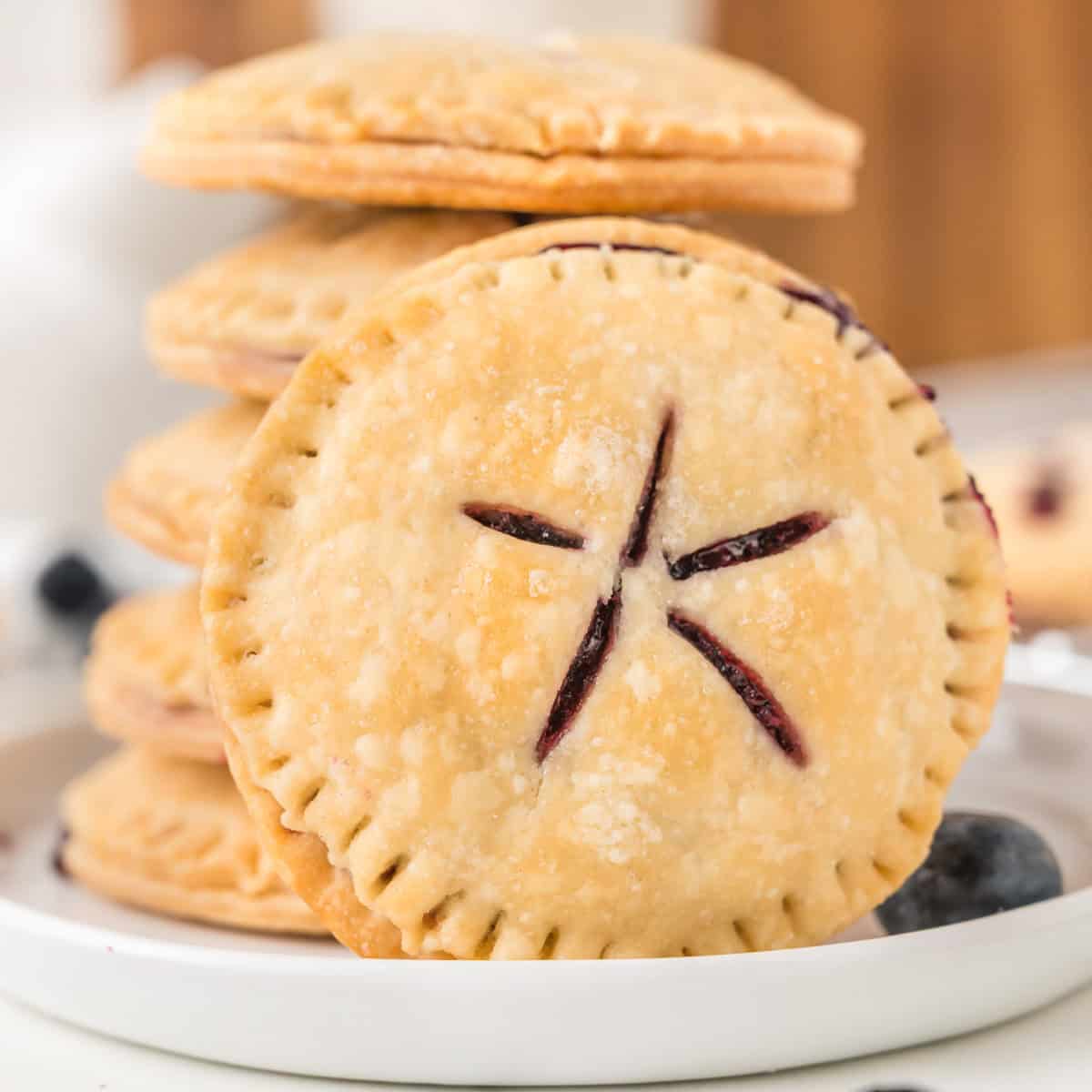 A stack of round, golden-brown hand pies with crimped edges and slit vents, displayed on a white plate with blueberries.