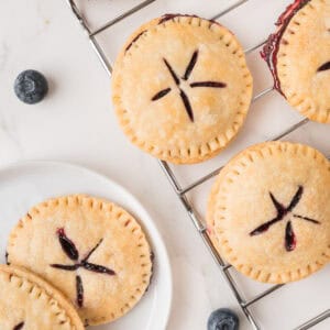 Round hand pies with golden crust and blueberry filling are arranged on a cooling rack and a white plate, with whole blueberries scattered nearby.