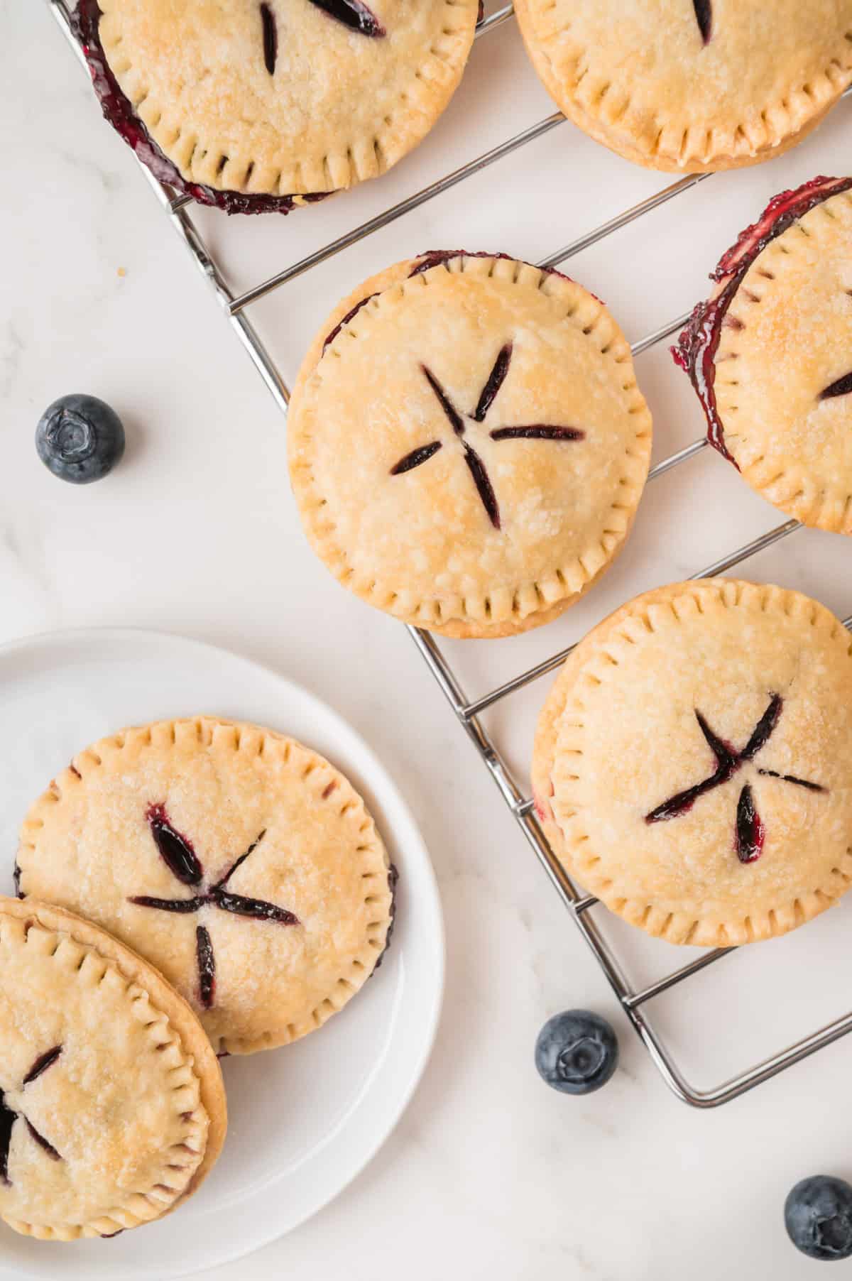 Round hand pies with golden crust and blueberry filling are arranged on a cooling rack and a white plate, with whole blueberries scattered nearby.