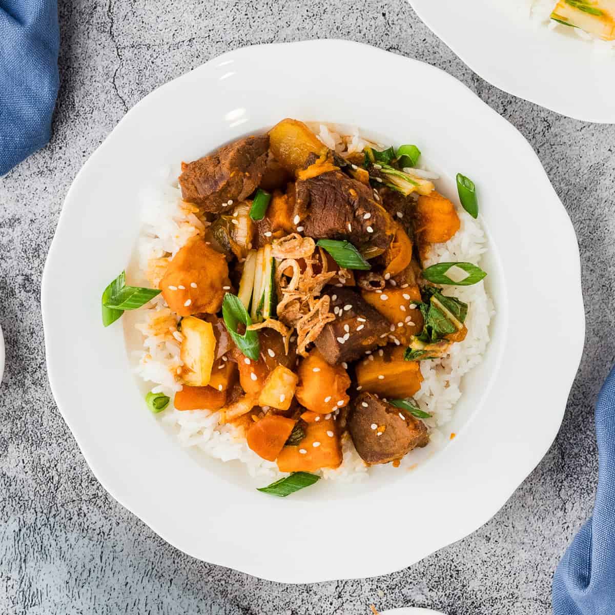 A white plate of rice topped with slow cooker Korean beef stew, packed with tender beef, vegetables, green onions, sesame seeds, and fried shallots.