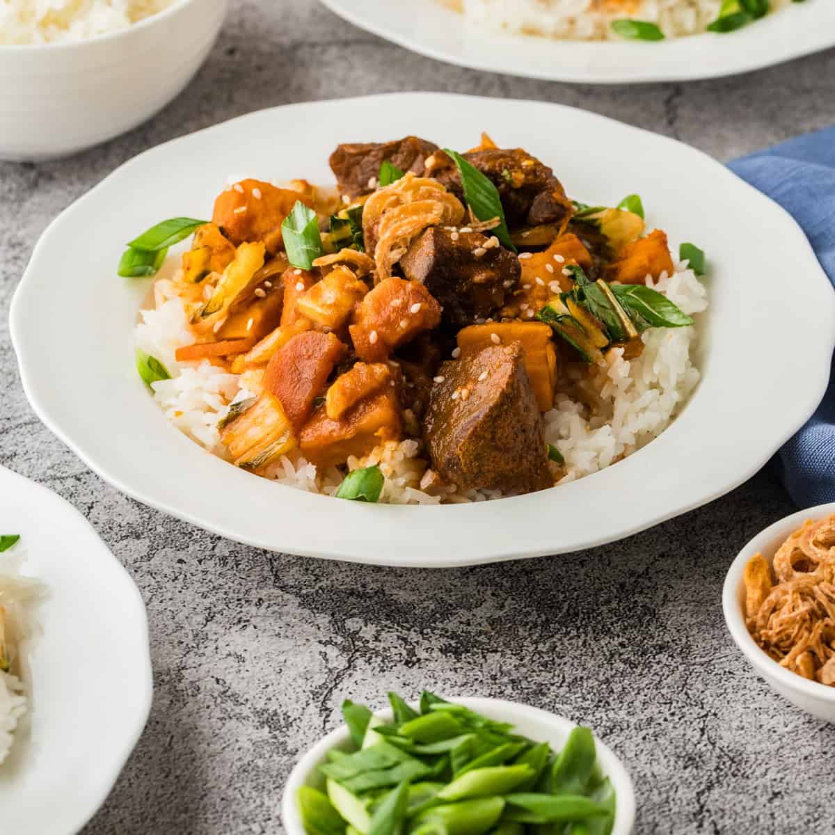 A plate of white rice topped with tender slow cooker Korean beef, vegetables, and garnished with chopped green onions, surrounded by small bowls of ingredients like sweet potato.