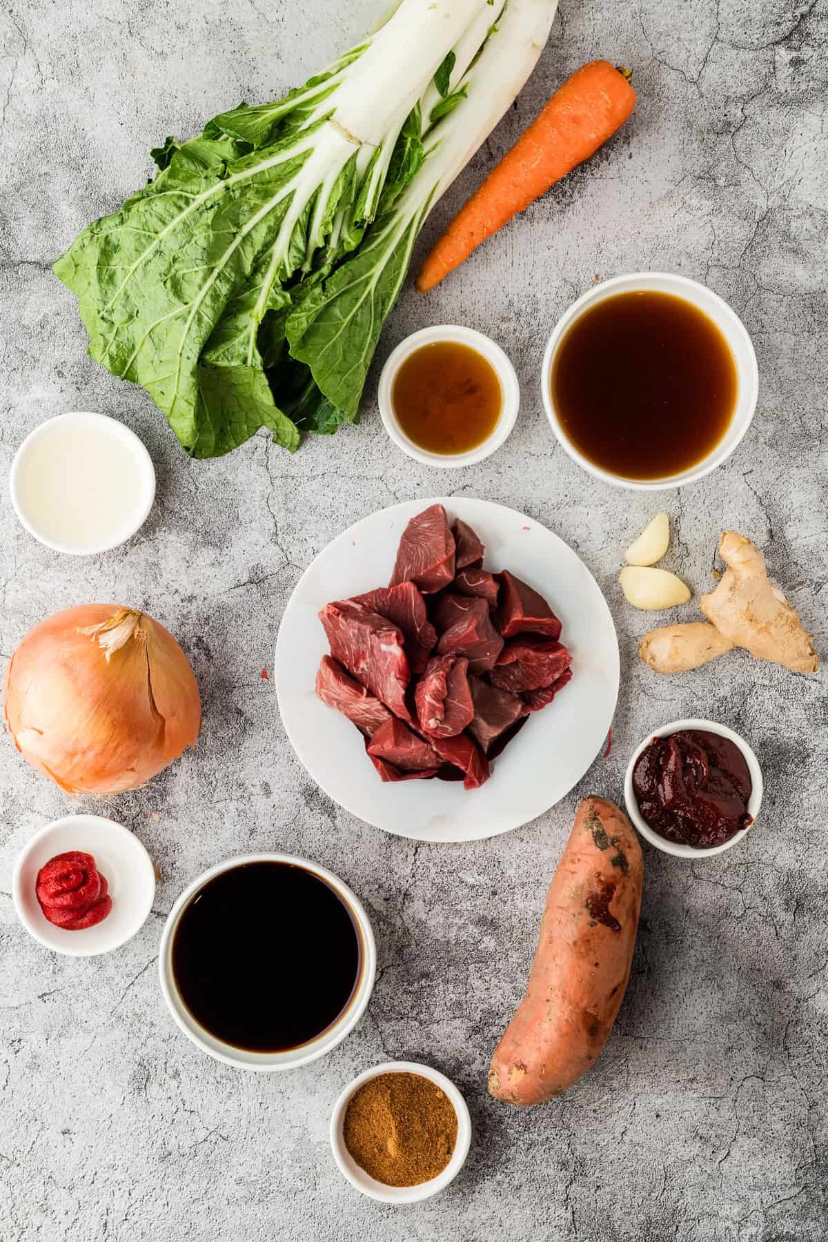 Assorted raw ingredients on a gray surface, including chunks of meat for slow cooker Korean beef, leafy greens, carrot, sweet potato, onion, garlic, ginger, sauces, broth, and seasonings in small bowls.