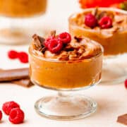 A glass dish filled with chocolate mousse, topped with chocolate pieces and fresh raspberries, sits on a light surface with more raspberries and chocolate nearby.