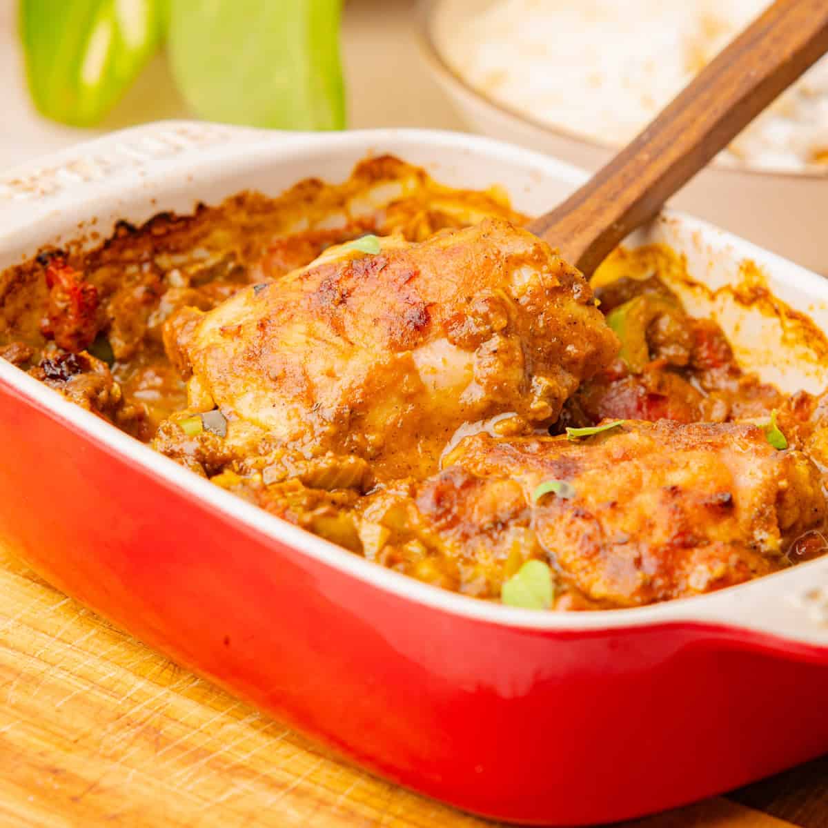 Baked chicken pieces in a red ceramic dish with a wooden spoon, covered in a golden brown sauce and herbs, placed on a wooden surface.