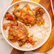 A white bowl filled with white rice and two pieces of stewed chicken with vegetables and sauce, placed on a wooden surface.