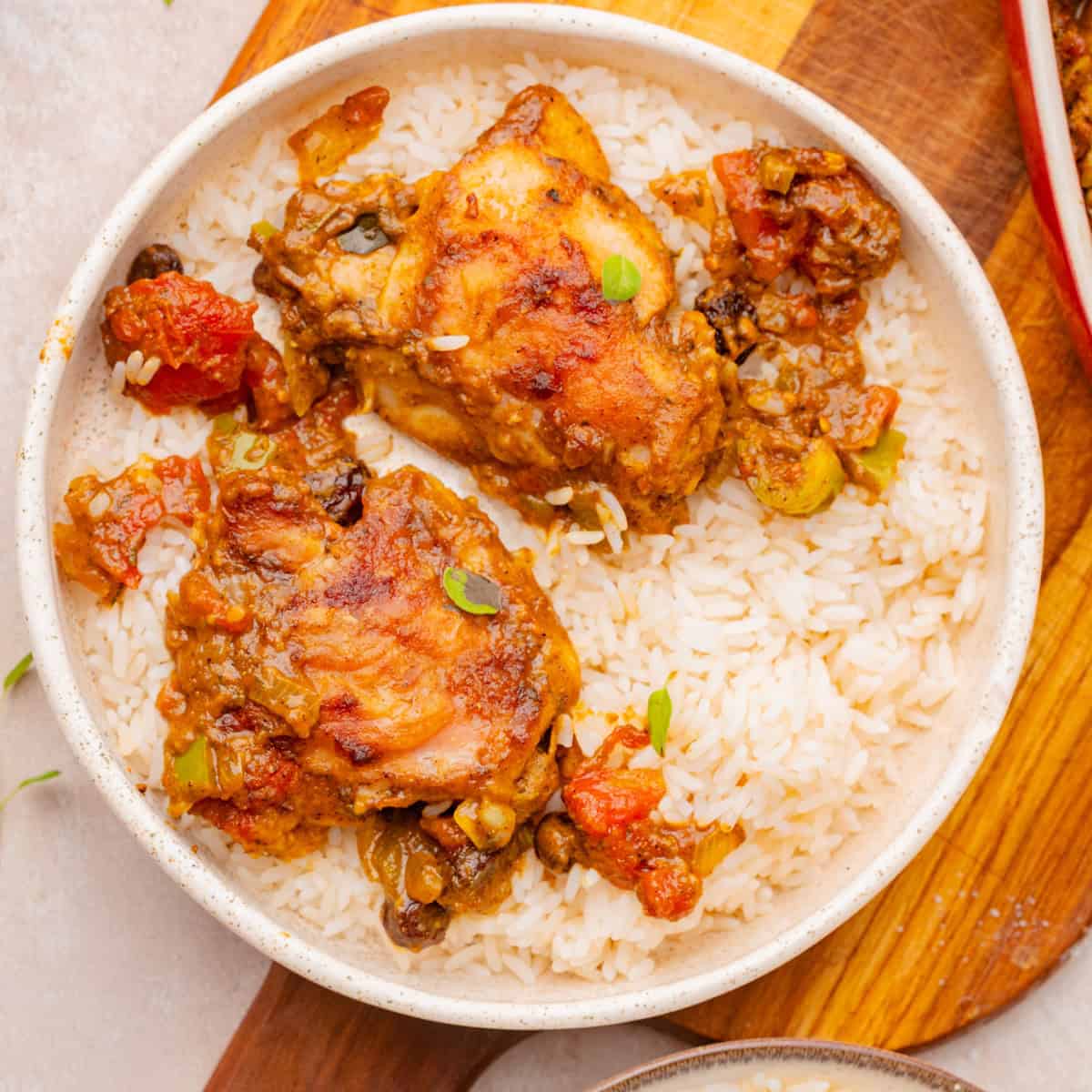 A white bowl filled with white rice and two pieces of stewed chicken with vegetables and sauce, placed on a wooden surface.