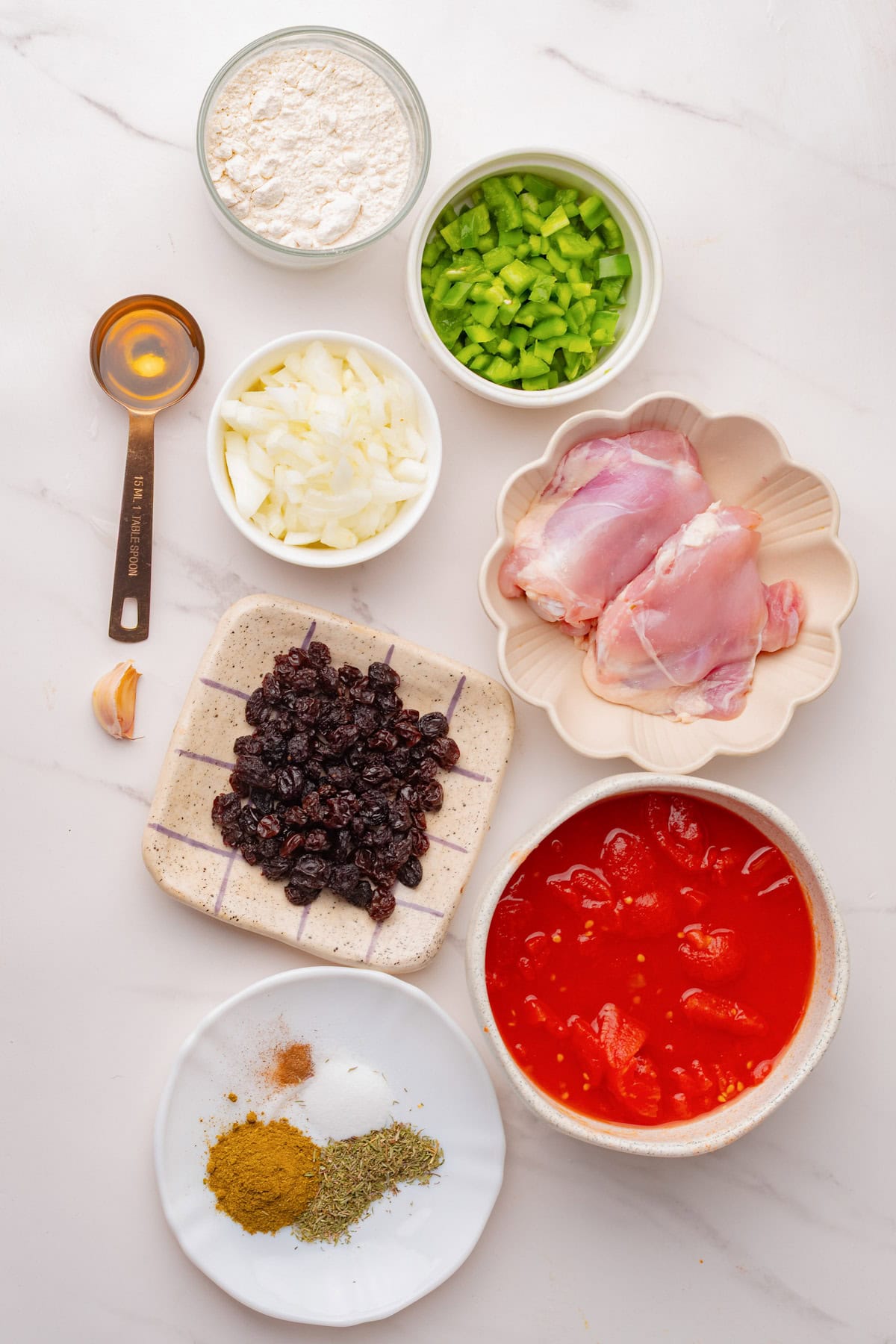 Ingredients for a Country Chicken Casserole arranged on a white surface, including chicken thighs, chopped onions, green peppers, diced tomatoes, raisins, flour, garlic, spices, and a tablespoon of honey.