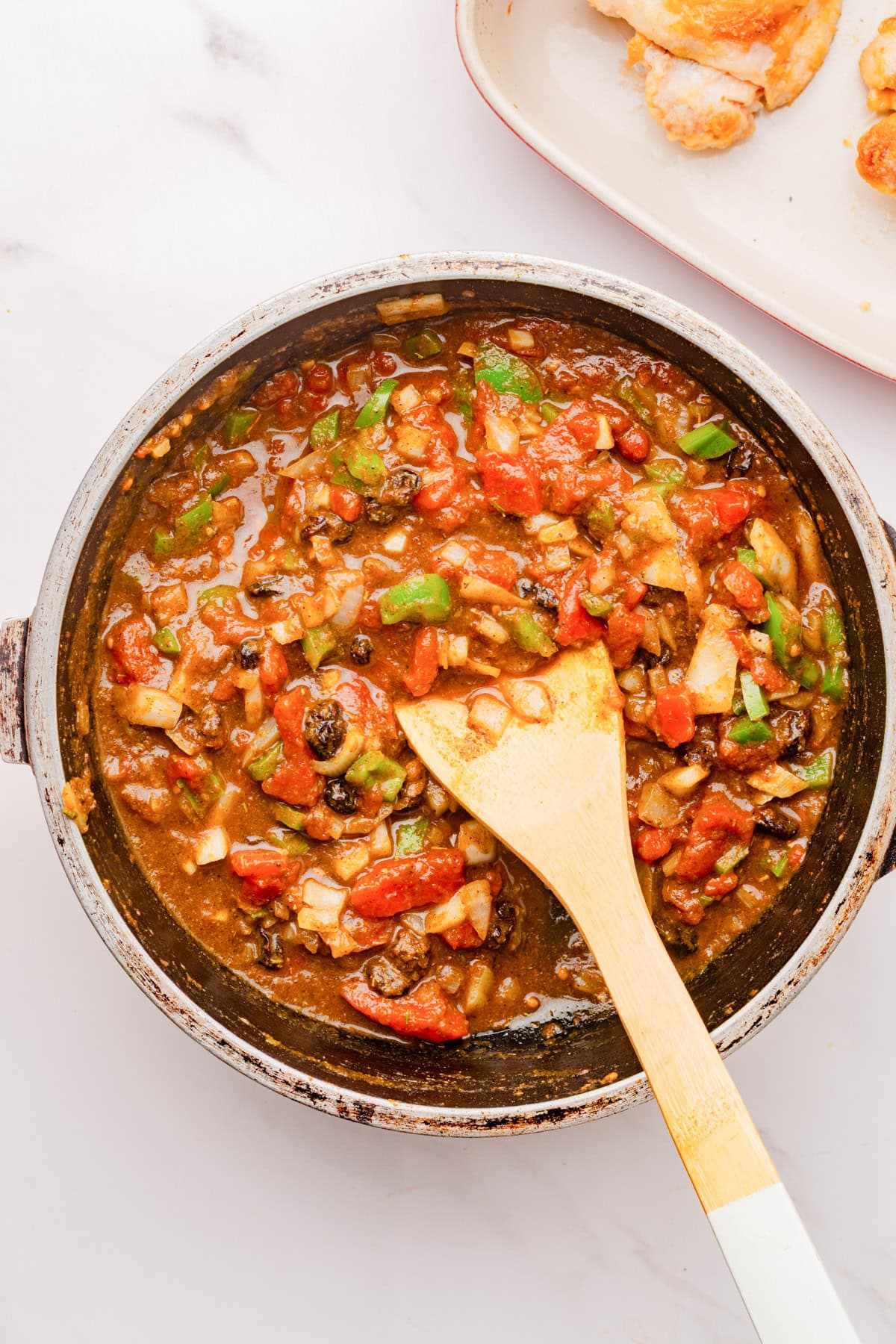 A skillet filled with a chunky tomato-based vegetable sauce is being stirred with a wooden spoon on a white countertop.