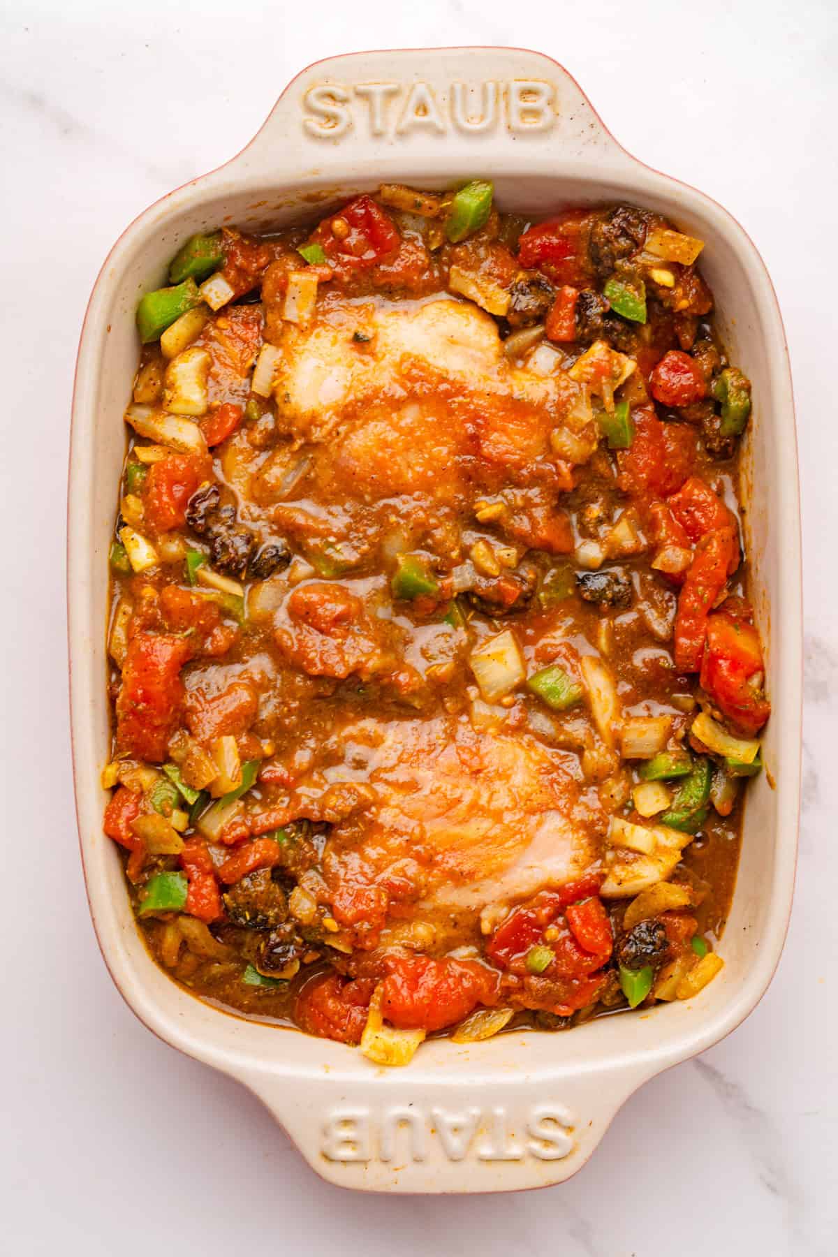 Baked chicken pieces in a ceramic Staub dish with chopped tomatoes, green bell peppers, onions, and a savory sauce.