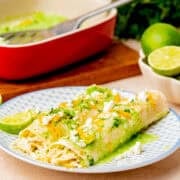 Two chicken enchiladas verdes topped with cheese, green sauce, and crumbled cheese are served on a patterned plate with lime halves in the background—an easy enchiladas dish perfect for any meal.