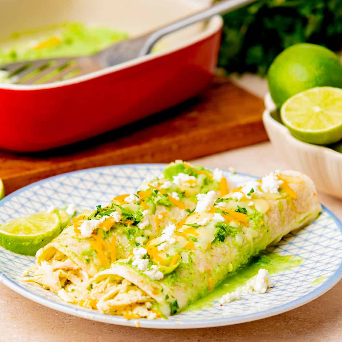 Two chicken enchiladas verdes topped with cheese, green sauce, and crumbled cheese are served on a patterned plate with lime halves in the background—an easy enchiladas dish perfect for any meal.