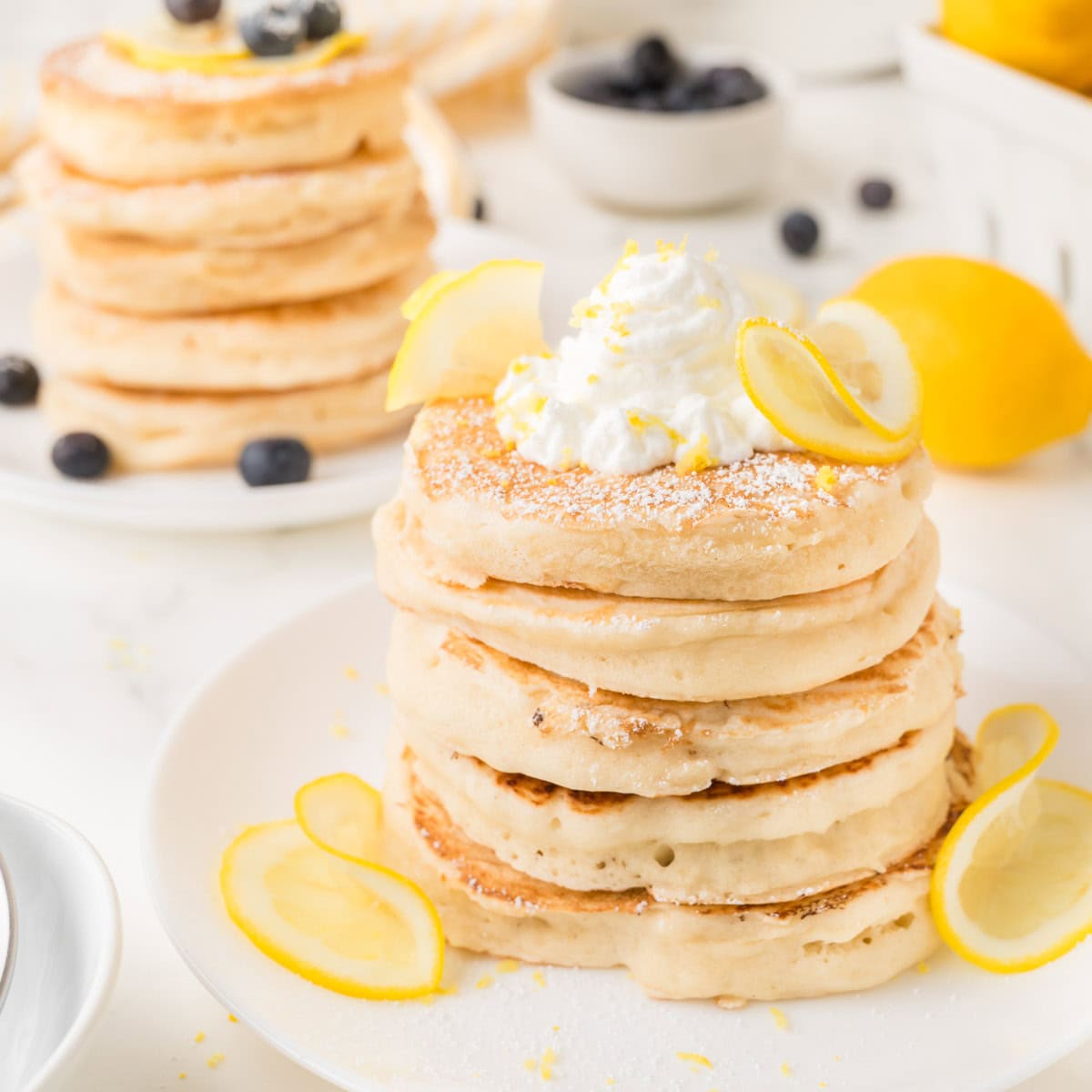 A stack of fluffy pancakes topped with whipped cream, lemon slices, and powdered sugar—perfect for those looking to use up lemons—with blueberries and extra lemons in the background.