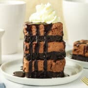 A tall slice of layered chocolate cheesecake with cookie crust, topped with whipped cream and drizzled with chocolate sauce, on a white plate.