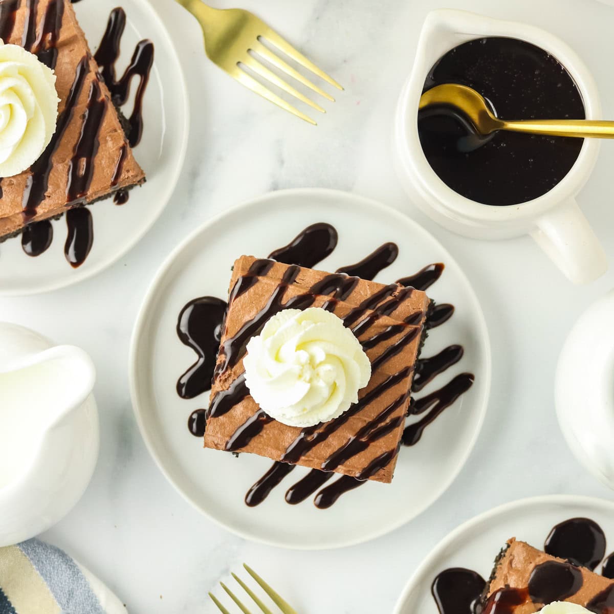 Square chocolate dessert with whipped cream and chocolate drizzle on a white plate, surrounded by gold forks, a pitcher of chocolate sauce, and a milk jug.