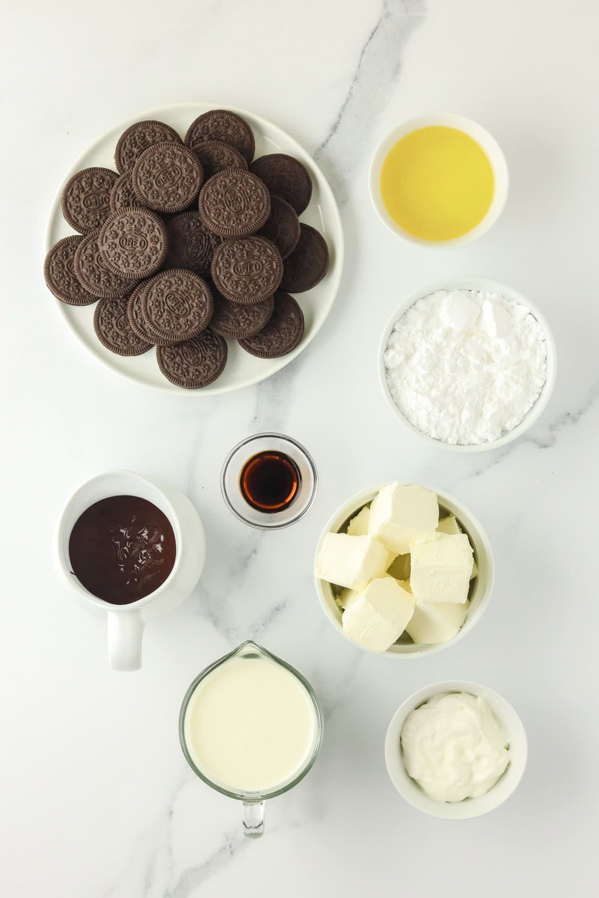 Ingredients for a dessert recipe arranged on a white surface: chocolate sandwich cookies, melted butter, powdered sugar, chocolate, vanilla, cream, cream cheese, and sour cream.