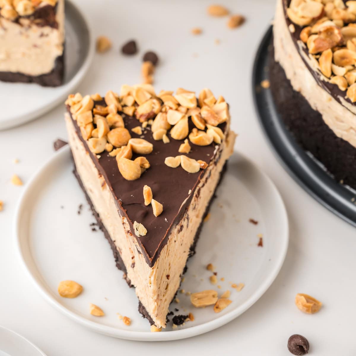 A slice of peanut butter cheesecake with a chocolate layer and chopped peanuts on top, served on a white plate.