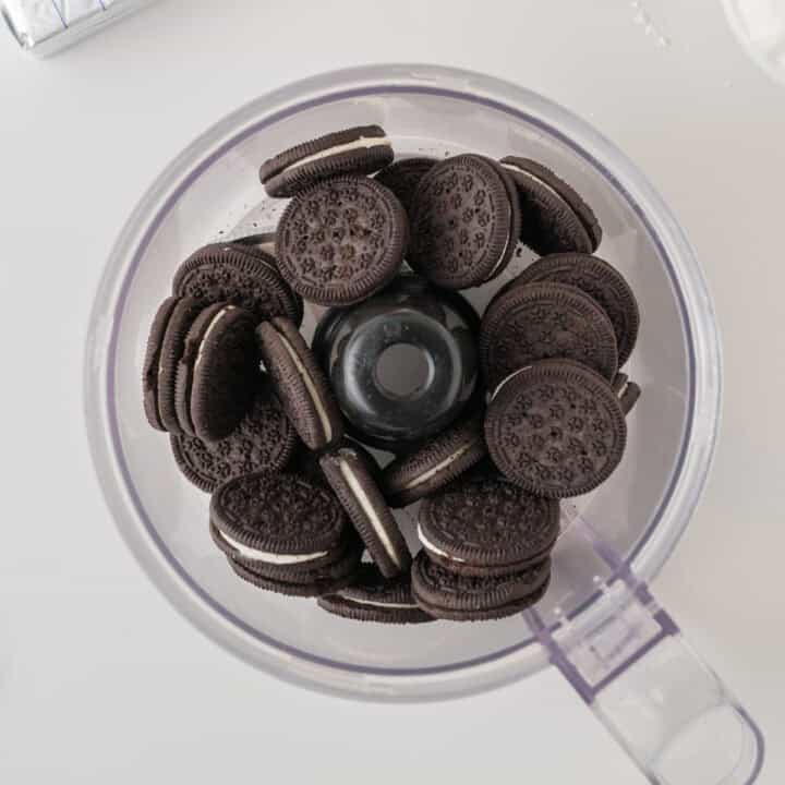 A clear food processor bowl filled with whole chocolate sandwich cookies on a white surface.