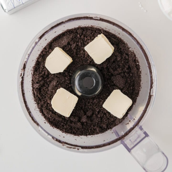 A food processor containing crushed chocolate cookies and four cubes of butter, viewed from above.