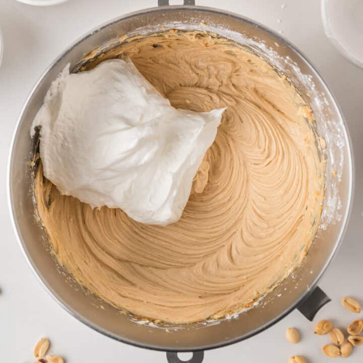 A mixing bowl containing creamy brown batter with a large dollop of white whipped topping about to be mixed in. Peanuts are scattered nearby.