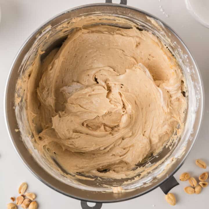 Mixing bowl filled with creamy peanut butter frosting, with some peanuts scattered nearby on a white surface.