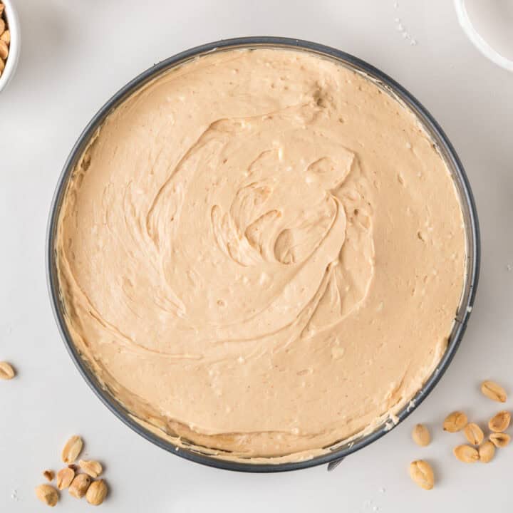 A round pan filled with creamy, light brown peanut butter cheesecake batter, surrounded by a few scattered peanuts on a white surface.