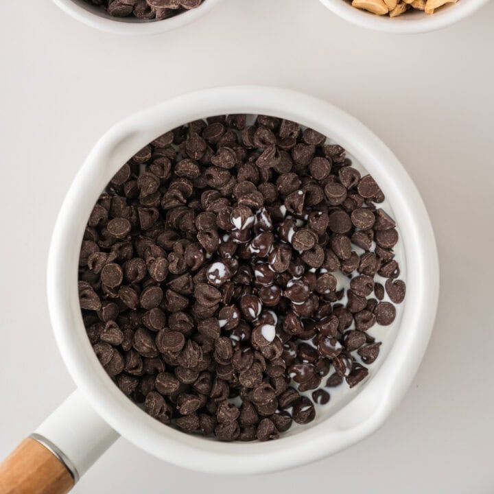 A white saucepan filled with chocolate chips and some melted chocolate, with parts of two bowls containing ingredients partially visible at the top.