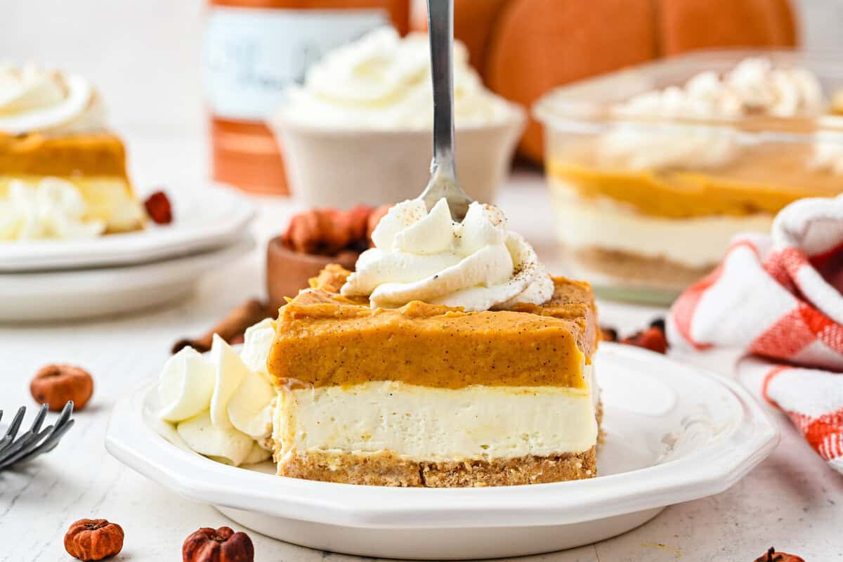 A fork is inserted into a slice of layered pumpkin cheesecake topped with whipped cream on a white plate, with more cheesecake and fall decor in the background.