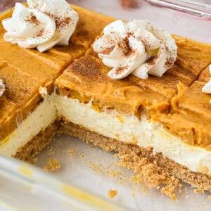 A glass dish containing layered pumpkin cheesecake bars with a graham cracker crust, topped with whipped cream and a sprinkle of cinnamon. A section has been cut out and removed.