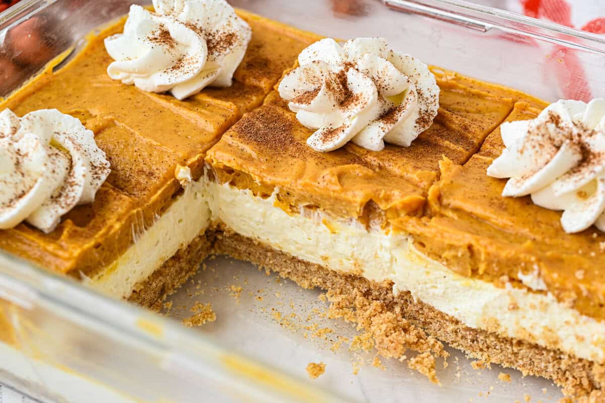 A glass dish containing layered pumpkin cheesecake bars with a graham cracker crust, topped with whipped cream and a sprinkle of cinnamon. A section has been cut out and removed.