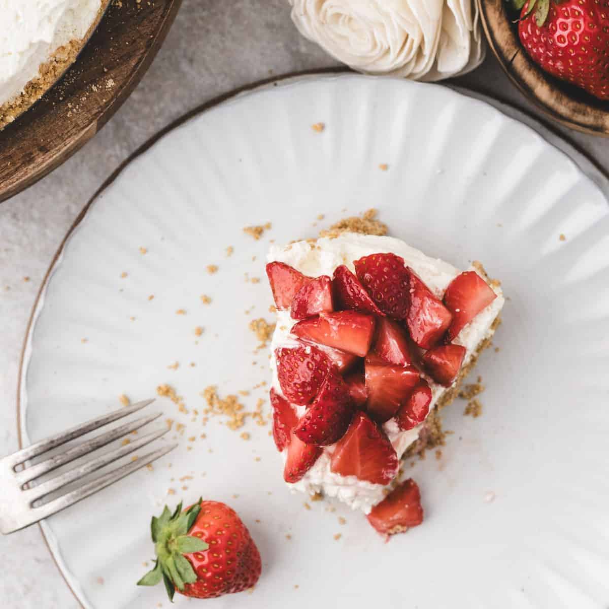 A slice of cheesecake topped with chopped strawberries sits on a white plate next to a fork, with a whole strawberry and flowers nearby.