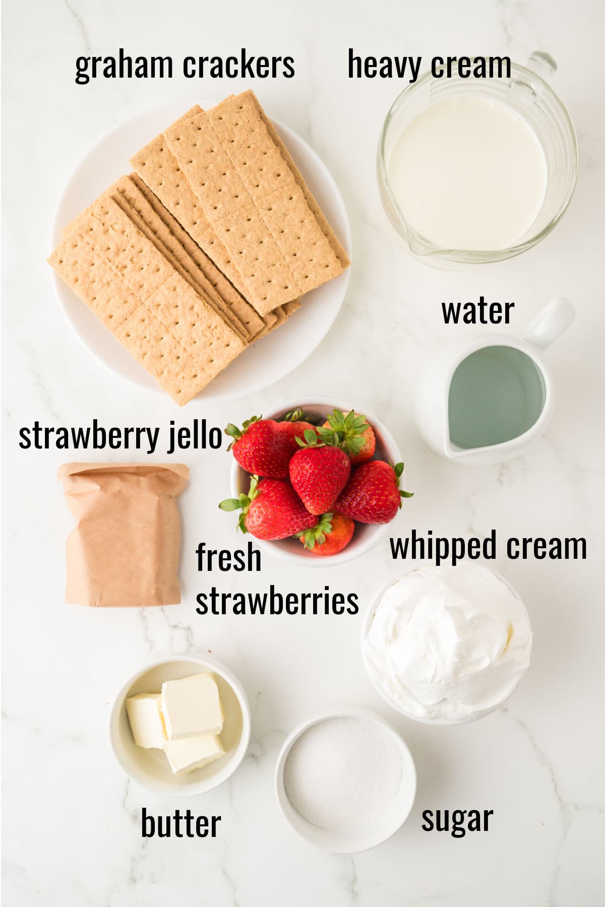 A top-down view of ingredients on a white surface: graham crackers, heavy cream, water, strawberry jello, fresh strawberries, whipped cream, butter, and sugar, each labeled.