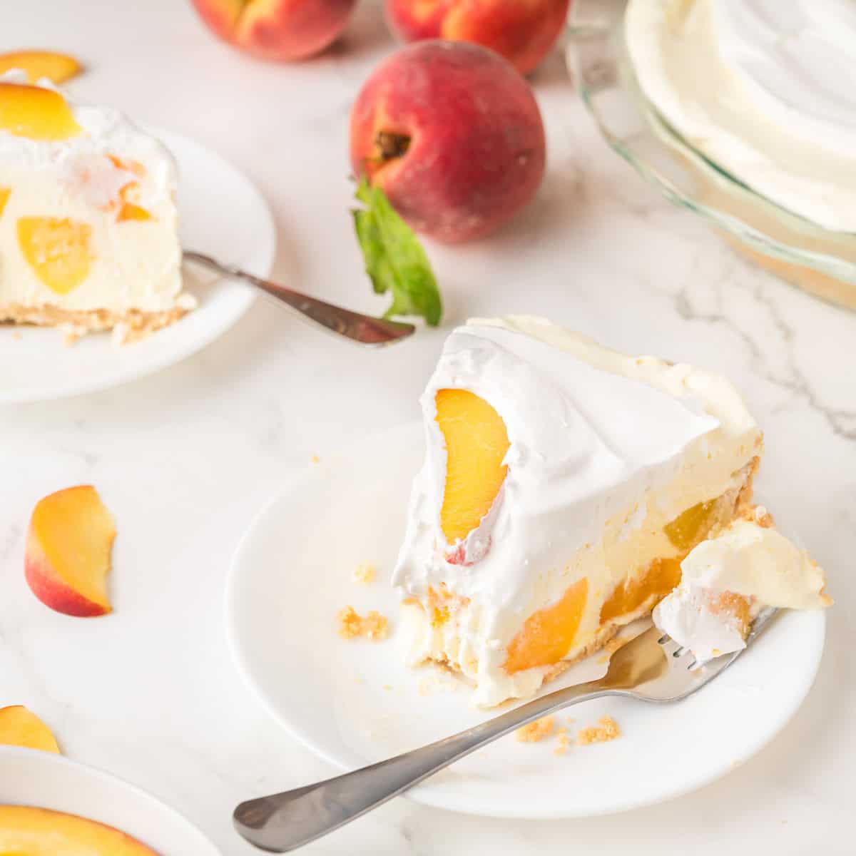 A slice of peach cream pie on a white plate with a fork, surrounded by fresh peaches and additional pie slices on a marble surface.