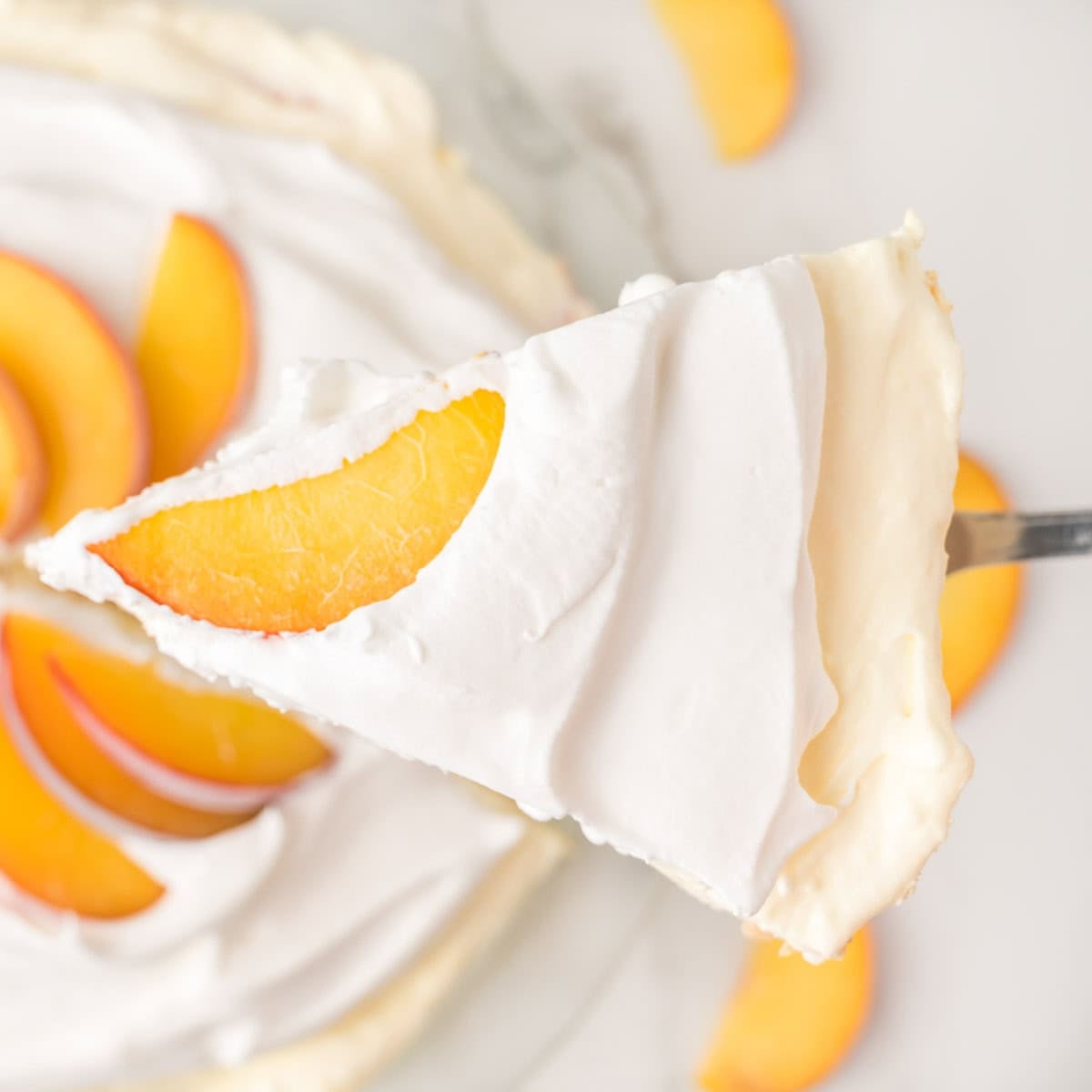 A close-up of a slice of cream pie topped with whipped cream and a peach slice, held above the rest of the pie on a serving utensil.