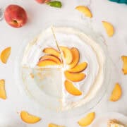 A cream pie topped with sliced peaches sits on a white surface; one slice is missing, and peach slices are scattered around the pie.