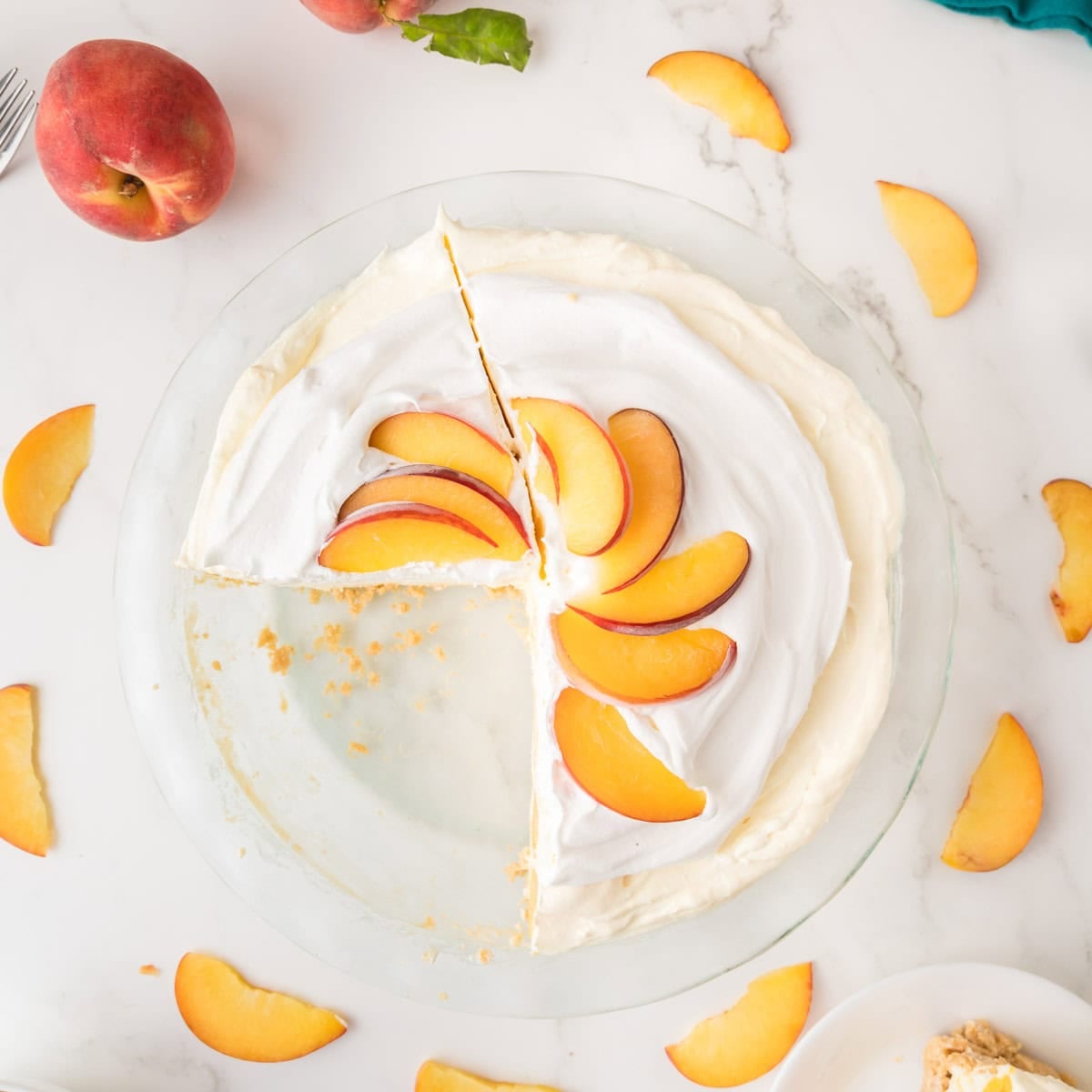 A cream pie topped with sliced peaches sits on a white surface; one slice is missing, and peach slices are scattered around the pie.