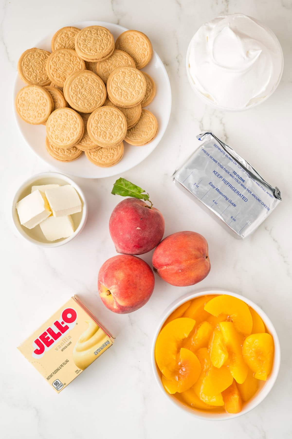 A flat lay of vanilla sandwich cookies, whipped topping, cream cheese, butter, fresh peaches, canned peach slices, and a box of vanilla pudding mix on a white surface.