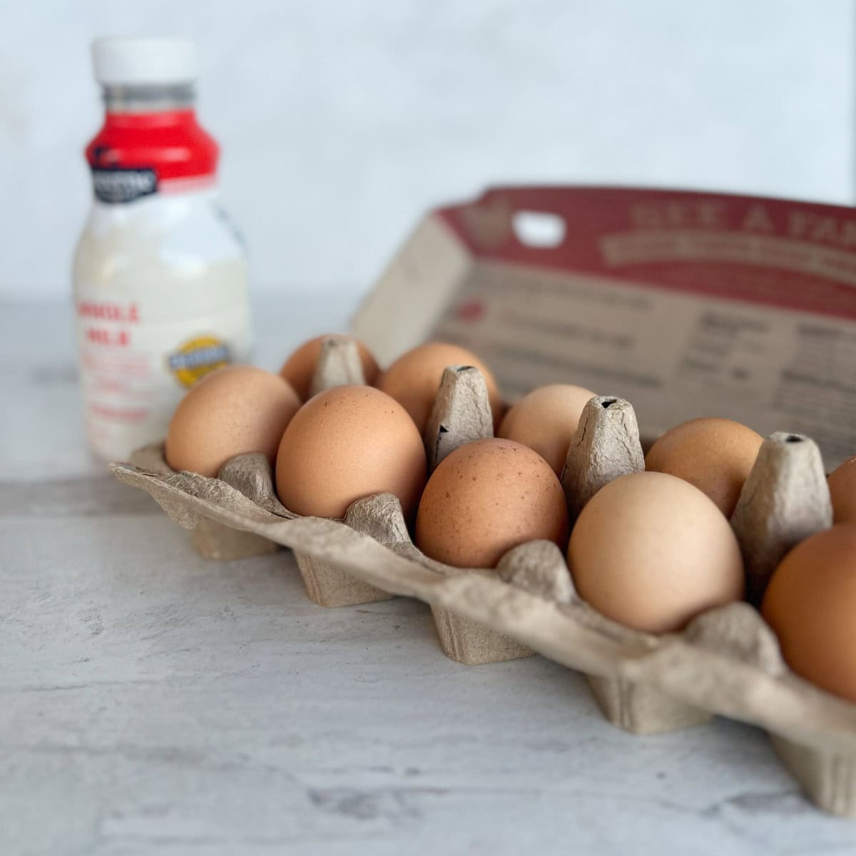 A carton of brown eggs sits on a light surface, perfect for making quiche custard, with a bottle of milk blurred in the background.