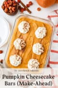 A glass dish of pumpkin cheesecake bars with six whipped cream dollops, surrounded by nuts, a pumpkin, and a red-striped towel on a white surface.