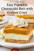 A slice of pumpkin cheesecake bar with graham crust, topped with whipped cream, on a white plate.