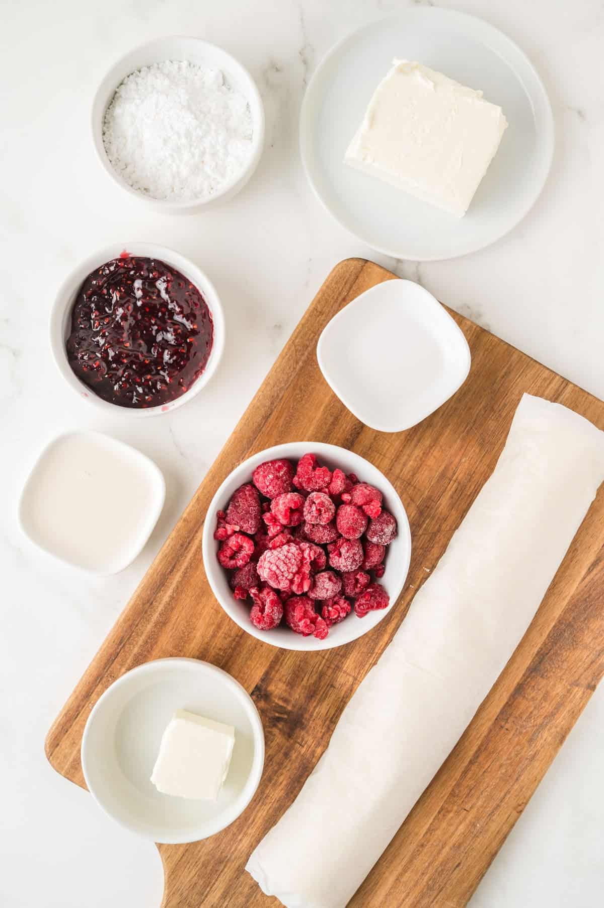 Overhead view of bowls containing powdered sugar, cream cheese, raspberry jam, frozen raspberries, butter, cream, and a rolled sheet of puff pastry on a wooden board.