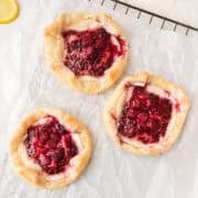 Three rustic berry galettes with a golden crust and red berry filling are arranged on parchment paper.