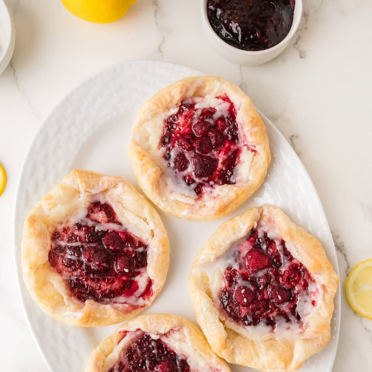 Four round pastries with berry filling and glaze are arranged on a white oval plate, with a bowl of berry jam and lemon slices nearby on a marble surface.