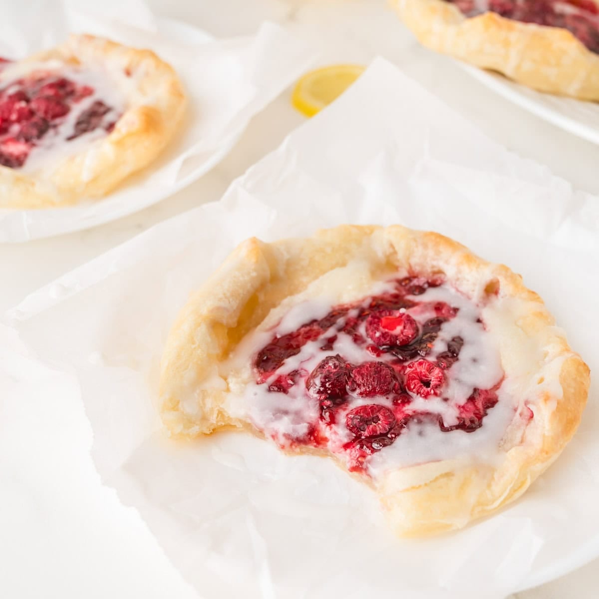 A raspberry pastry with icing on parchment paper, showing a bite taken out of it. Other similar pastries are in the background.