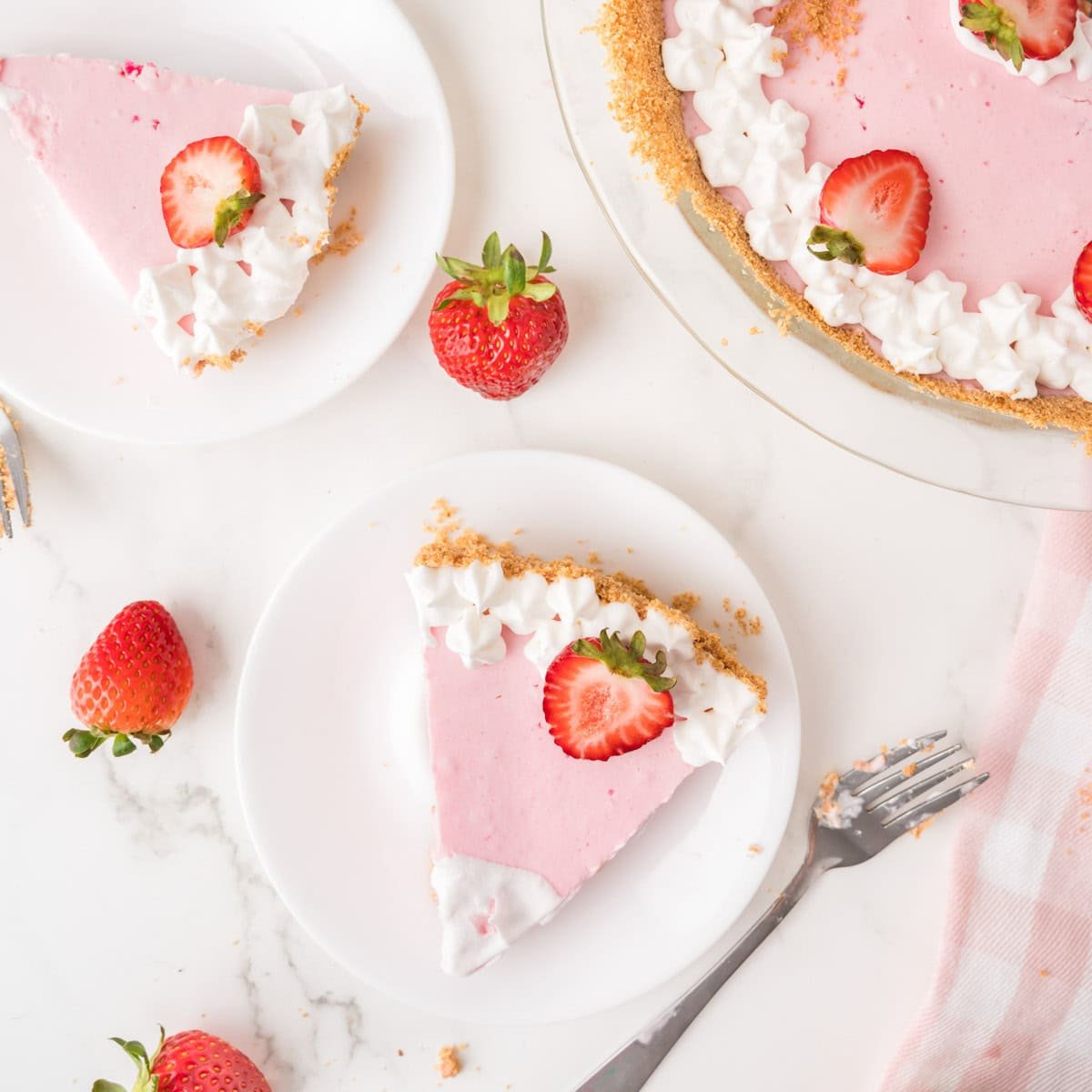 A slice of strawberry cream pie with whipped cream and a fresh strawberry sits on a plate, next to the rest of the pie and scattered strawberries on a marble surface.
