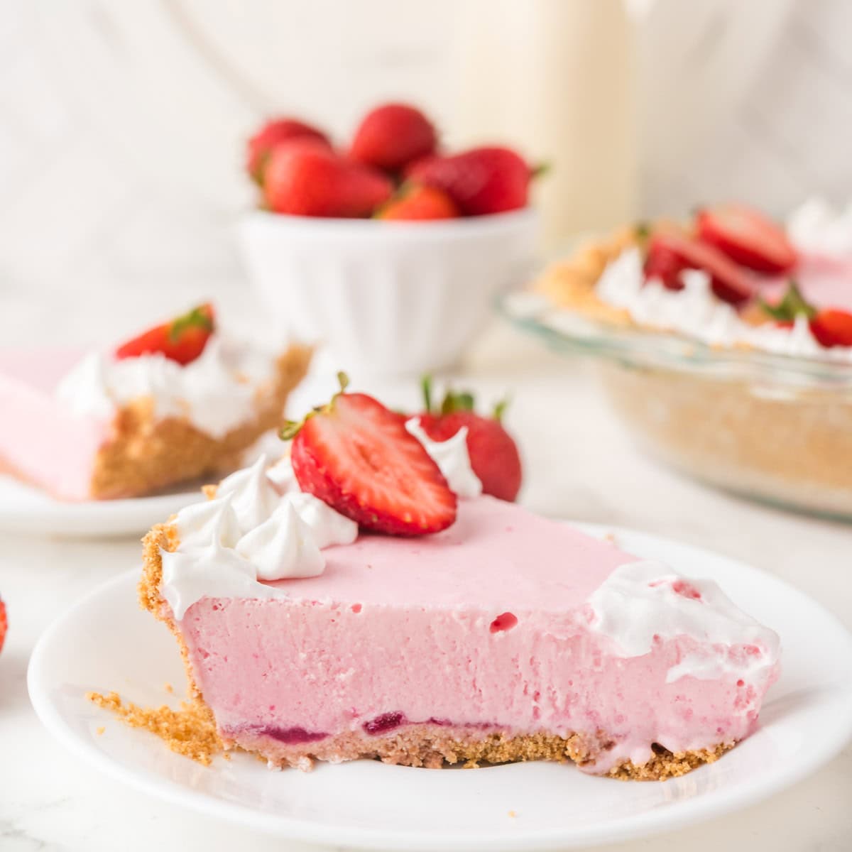 A slice of pink strawberry cream pie with a graham cracker crust, whipped cream, and fresh strawberry slices on a white plate. Whole strawberries and more pie are in the background.