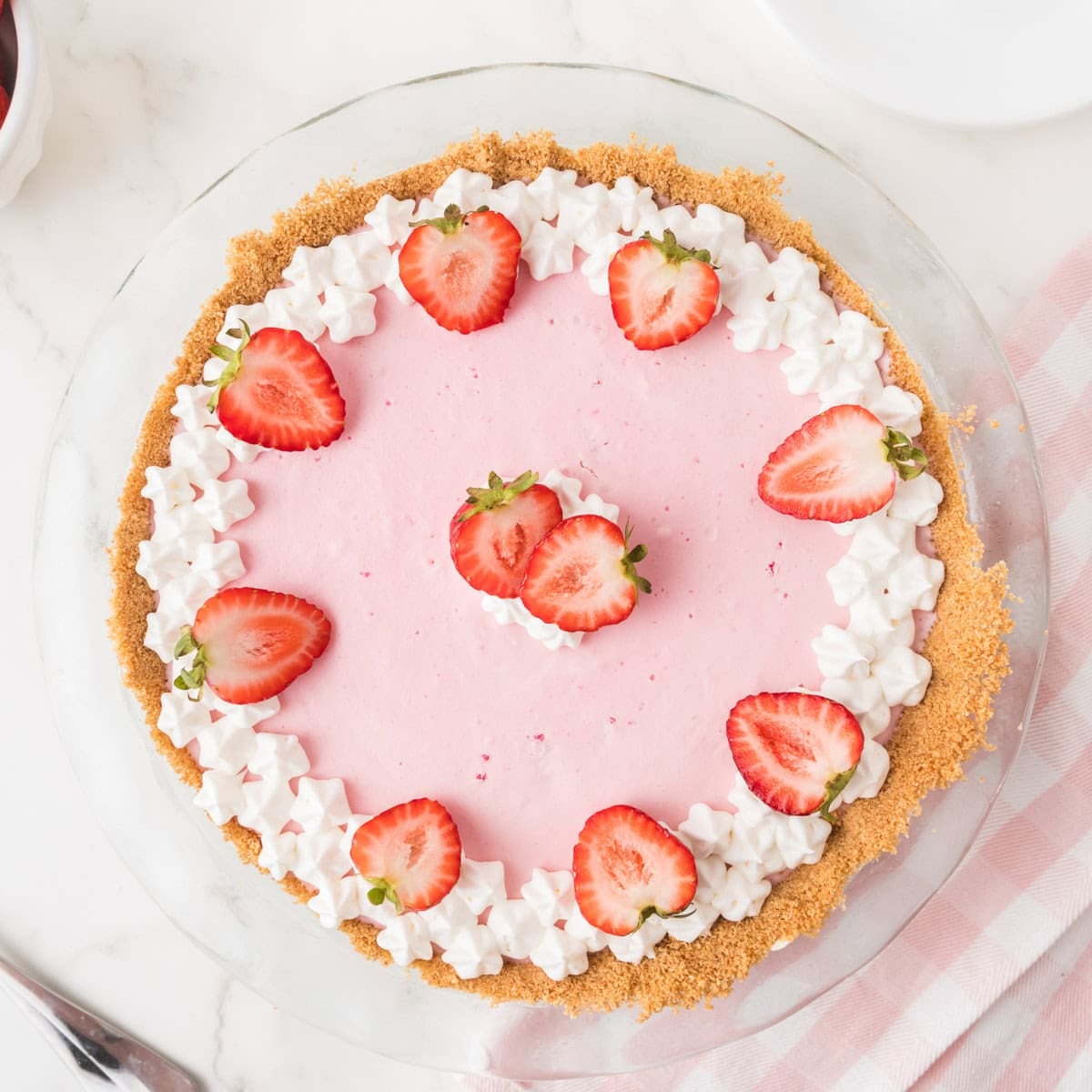 A strawberry cream pie with a graham cracker crust, topped with whipped cream and halved strawberries, on a white surface.